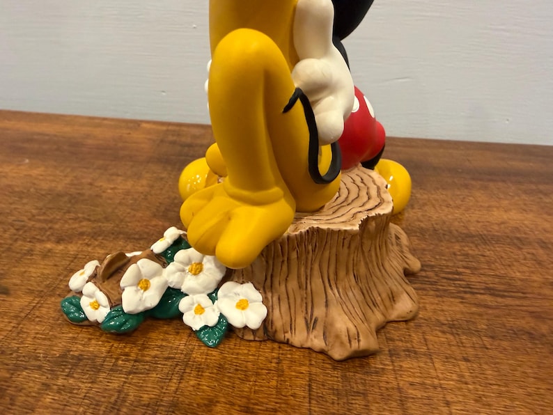 K&ouml;nnte beinhalten: Eine Keramikfigur mit Micky Maus und Pluto. Micky tr&auml;gt wei&szlig;e Handschuhe, rote Shorts und schwarze Schuhe. Pluto hat gelbe Beine und F&uuml;&szlig;e. Die Figur steht auf einem Baumstumpf mit Blumen.