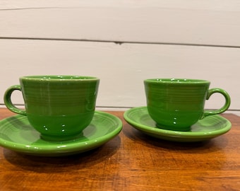 May include: Two matching bright green ceramic tea cups and saucers. Each cup features a rounded body with horizontal ringed detailing and a curved handle. The matching saucers have a smooth, circular design with a slightly raised center.