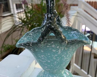 May include: A vintage ceramic basket with a turquoise speckled glaze. The basket has a pedestal base and a ruffled rim. The handle is dark green with white speckles. A decorative piece for displaying flowers or other items.