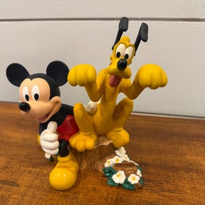 K&ouml;nnte beinhalten: Eine farbenfrohe Figur mit Micky Maus und Pluto. Micky tr&auml;gt sein klassisches Outfit, und Pluto ist in einer spielerischen Pose. Der Sockel enth&auml;lt ein Blumenarrangement. Ein bezauberndes Sammlerst&uuml;ck.