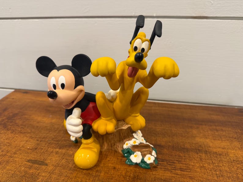 K&ouml;nnte beinhalten: Eine farbenfrohe Figur mit Micky Maus und Pluto. Micky tr&auml;gt sein klassisches Outfit, und Pluto ist in einer spielerischen Pose. Der Sockel enth&auml;lt ein Blumenarrangement. Ein bezauberndes Sammlerst&uuml;ck.