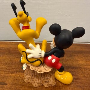 K&ouml;nnte beinhalten: Eine Figur mit Micky Maus und Pluto. Micky tr&auml;gt ein schwarzes Hemd, rote Shorts und gelbe Schuhe, w&auml;hrend Pluto gelb mit schwarzen Ohren und einem roten Halsband ist. Der Sockel ist ein Baumstumpf.