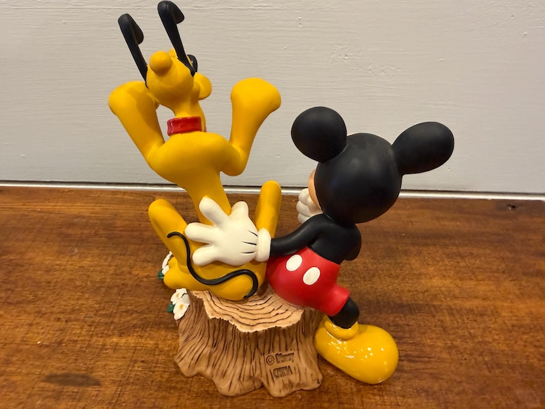 K&ouml;nnte beinhalten: Eine Figur mit Micky Maus und Pluto. Micky tr&auml;gt ein schwarzes Hemd, rote Shorts und gelbe Schuhe, w&auml;hrend Pluto gelb mit schwarzen Ohren und einem roten Halsband ist. Der Sockel ist ein Baumstumpf.