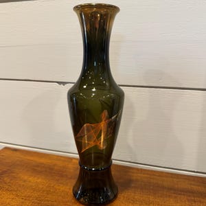 Puede incluir: Un jarrón alto de vidrio verde oscuro con una base acampanada y un cuello estrecho. El borde tiene un acabado dorado. Un diseño abstracto naranja y dorado está en la parte delantera del jarrón. El jarrón mide aproximadamente 25 cm de alto.