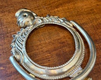 Könnte beinhalten: Verzierter, goldfarbener Metallrahmen mit kreisförmiger Öffnung. Der Rahmen weist detaillierte Reliefschnitzereien auf, darunter ein Blumenmotiv und eine dekorative Bordüre. Er hat eine gebogene Form und einen kleinen, quadratischen Sockel. Das Design ist vintage und dekorativ.