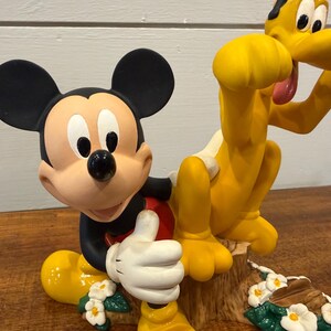K&ouml;nnte beinhalten: Eine Figur mit Micky Maus und Pluto. Micky ist in einer klassischen Pose mit erhobenem Daumen, tr&auml;gt schwarze Shorts, ein rotes Hemd und wei&szlig;e Handschuhe. Pluto ist gelb mit einem wei&szlig;en Fleck und einer rosa Zunge. Die Basis hat Blumen.