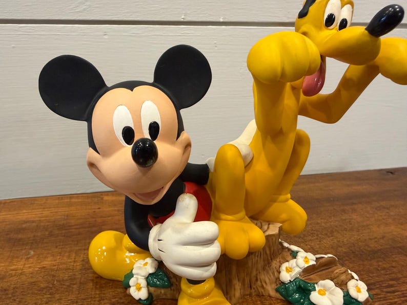 K&ouml;nnte beinhalten: Eine Figur mit Micky Maus und Pluto. Micky ist in einer klassischen Pose mit erhobenem Daumen, tr&auml;gt schwarze Shorts, ein rotes Hemd und wei&szlig;e Handschuhe. Pluto ist gelb mit einem wei&szlig;en Fleck und einer rosa Zunge. Die Basis hat Blumen.