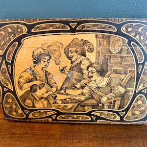 May include: Vintage wooden box with a black and beige Art Nouveau design. The box features a sepia-toned illustration of three people in period clothing, seated around a table, with one smoking a pipe. The box is rectangular with a curved top.