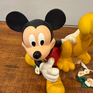 K&ouml;nnte beinhalten: Eine Mickey Mouse-Figur mit schwarzen Ohren, einem hellbraunen Gesicht und einem roten Hemd. Mickey hebt den Daumen. Pluto ist ebenfalls in der Figur, mit gelbem Fell und wei&szlig;en Handschuhen. Die Figur ist mit wei&szlig;en Blumen und gr&uuml;nen Bl&auml;ttern verziert.