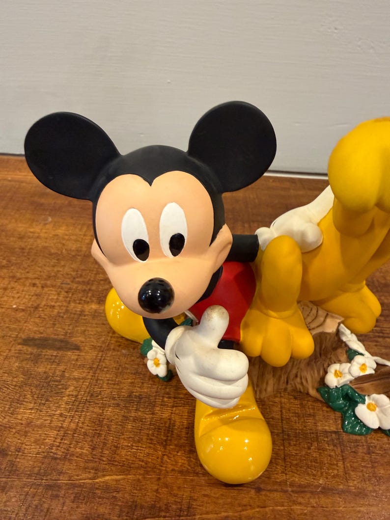 K&ouml;nnte beinhalten: Eine Mickey Mouse-Figur mit schwarzen Ohren, einem hellbraunen Gesicht und einem roten Hemd. Mickey hebt den Daumen. Pluto ist ebenfalls in der Figur, mit gelbem Fell und wei&szlig;en Handschuhen. Die Figur ist mit wei&szlig;en Blumen und gr&uuml;nen Bl&auml;ttern verziert.