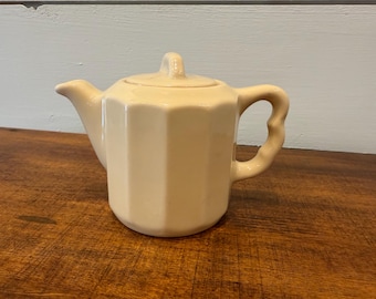 May include: Cream-colored ceramic teapot with a lid and a curved handle. The teapot has a faceted body and a spout. The handle is curved and the lid has a small handle. The teapot is sitting on a wooden surface.