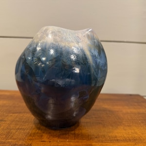 May include: A handcrafted, organic-shaped ceramic vase with a unique crystalline glaze. The vase transitions from a dark, almost black base to shades of blue and white at the top. The vase is displayed on a wooden surface.