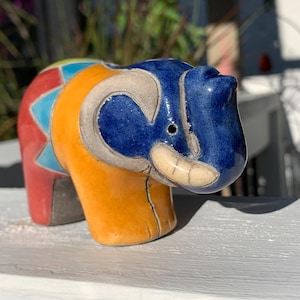 May include: A colorful ceramic elephant figurine. The elephant is painted with a blue head, orange body, and red and blue accents. The figurine has a smiling expression and is displayed on a white surface.