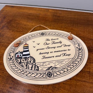 May include: Oval ceramic wall plaque featuring a lighthouse illustration and the text "The love in Our Family grows Strong and Deep leaving us memories to Treasure and Keep". It has a rope for hanging.