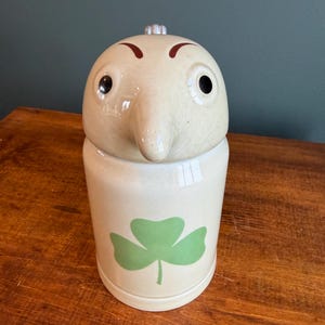 Peut inclure: Un pot à biscuits en céramique avec un design de visage fantaisiste. Le pot est blanc cassé avec un trèfle vert sur le devant. Le couvercle a la forme d'une tête avec un long nez, des yeux noirs et des sourcils bruns.
