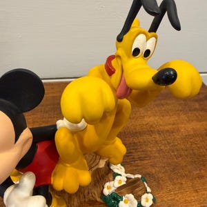 K&ouml;nnte beinhalten: Eine farbenfrohe Figur mit Micky Maus und Pluto. Micky tr&auml;gt rote Shorts und wei&szlig;e Handschuhe. Pluto ist gelb mit schwarzen Ohren und einem roten Halsband. Der Sockel hat wei&szlig;e Blumen und einen Baumstamm.