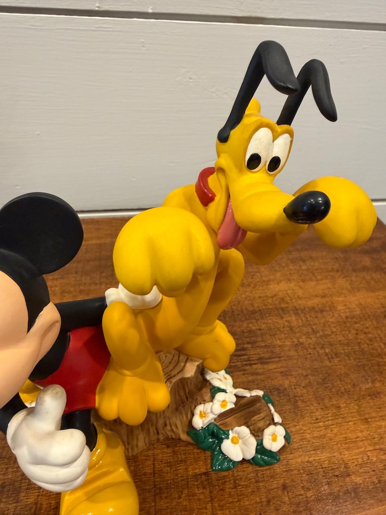 K&ouml;nnte beinhalten: Eine farbenfrohe Figur mit Micky Maus und Pluto. Micky tr&auml;gt rote Shorts und wei&szlig;e Handschuhe. Pluto ist gelb mit schwarzen Ohren und einem roten Halsband. Der Sockel hat wei&szlig;e Blumen und einen Baumstamm.