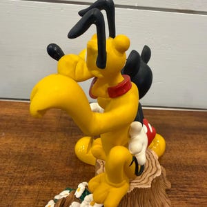 K&ouml;nnte beinhalten: Eine gelbe Pluto-Figur mit schwarzen Ohren und rotem Halsband, posiert mit Micky Maus. Die Figur steht auf einem Baumstumpf-Sockel mit wei&szlig;en Blumen und gr&uuml;nen Bl&auml;ttern. Ein Sammlerst&uuml;ck.