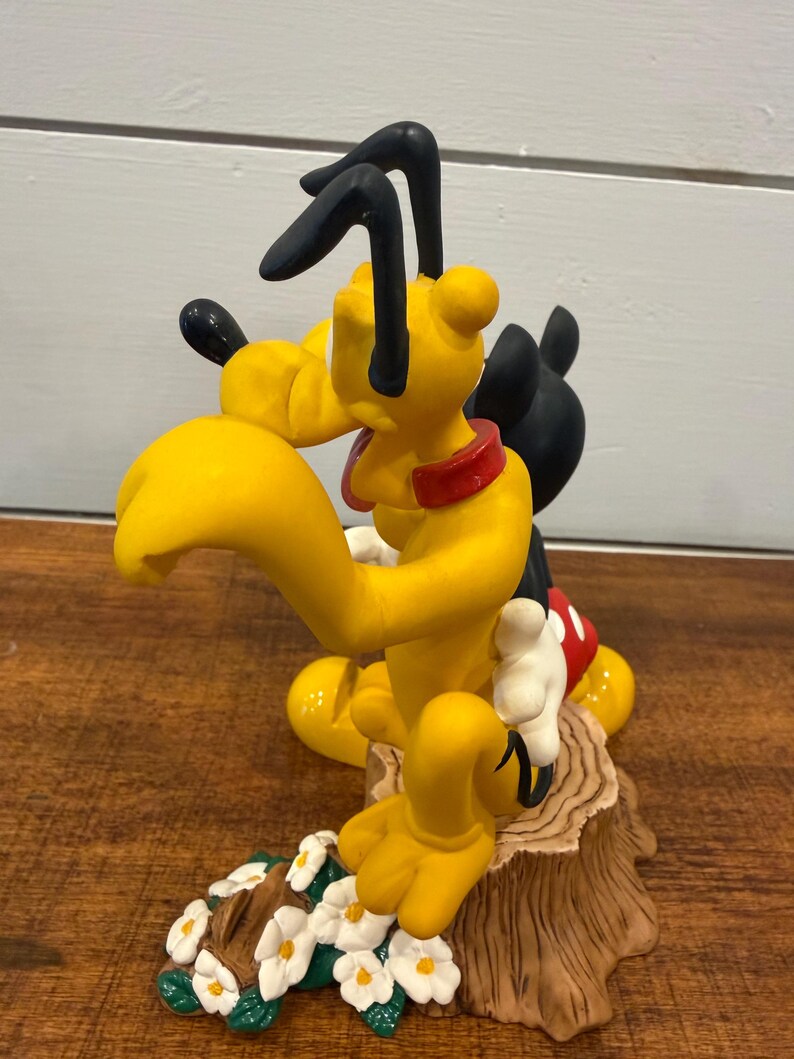 K&ouml;nnte beinhalten: Eine gelbe Pluto-Figur mit schwarzen Ohren und rotem Halsband, posiert mit Micky Maus. Die Figur steht auf einem Baumstumpf-Sockel mit wei&szlig;en Blumen und gr&uuml;nen Bl&auml;ttern. Ein Sammlerst&uuml;ck.