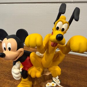 K&ouml;nnte beinhalten: Eine farbenfrohe Figur mit Micky Maus und Pluto. Micky tr&auml;gt sein klassisches Outfit, und Pluto ist leuchtend gelb. Die Figur steht auf einer Holzoberfl&auml;che und hat wei&szlig;e Blumen am Sockel.