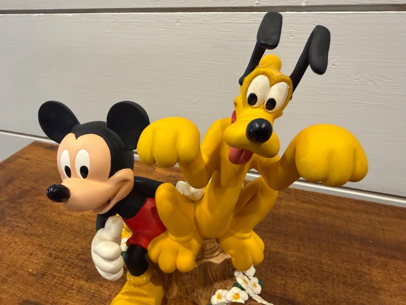 K&ouml;nnte beinhalten: Eine farbenfrohe Figur mit Micky Maus und Pluto. Micky tr&auml;gt sein klassisches Outfit, und Pluto ist leuchtend gelb. Die Figur steht auf einer Holzoberfl&auml;che und hat wei&szlig;e Blumen am Sockel.