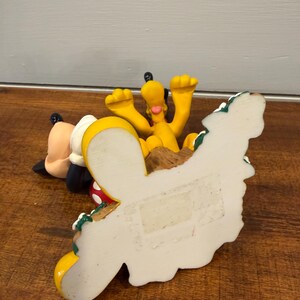 K&ouml;nnte beinhalten: Eine gelbe Pluto-Figur mit erhobenen Armen, neben einer Mickey Mouse-Figur. Die Figuren stehen auf einer Holzoberfl&auml;che. Der Sockel der Pluto-Figur ist wei&szlig; mit einem dekorativen Rand.