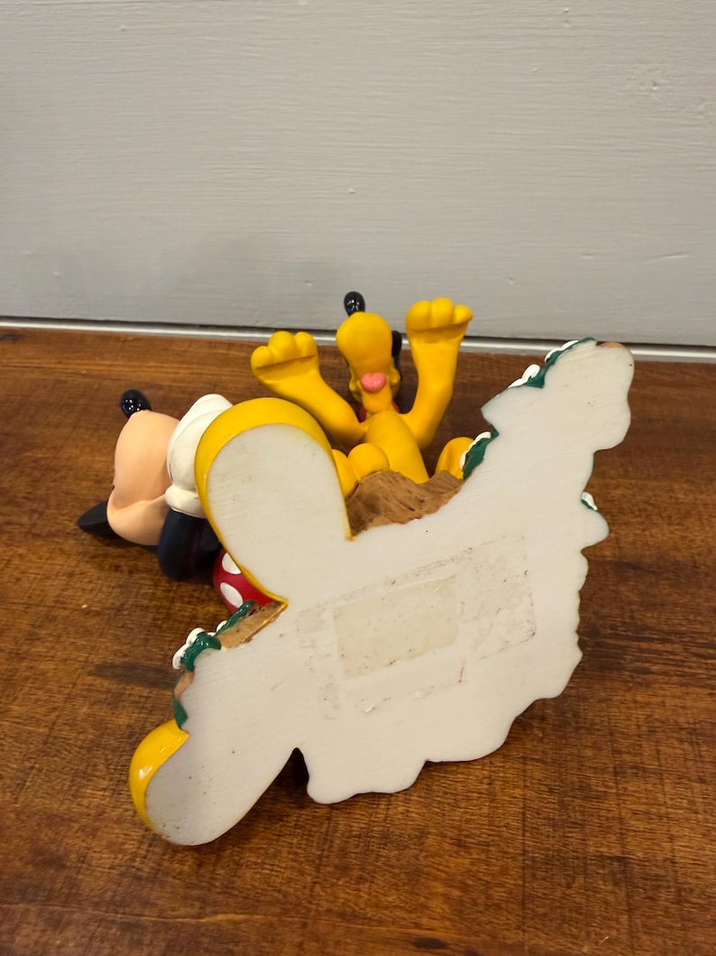 K&ouml;nnte beinhalten: Eine gelbe Pluto-Figur mit erhobenen Armen, neben einer Mickey Mouse-Figur. Die Figuren stehen auf einer Holzoberfl&auml;che. Der Sockel der Pluto-Figur ist wei&szlig; mit einem dekorativen Rand.