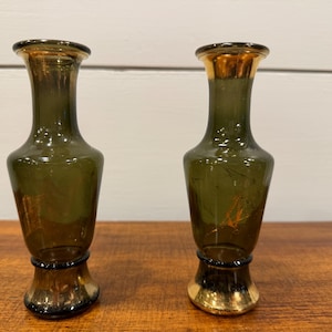 Puede incluir: Dos jarrones de vidrio verde oliva con bordes y bases doradas. Los jarrones tienen una forma delgada y alargada con una parte superior acampanada y una banda dorada decorativa cerca de la base. Los jarrones se exhiben sobre una superficie de madera.