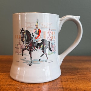 May include: White ceramic beer stein with a handle, featuring a colorful illustration of a Life Guard on horseback. The illustration includes the text "Trooper" and "Life Guards". The stein is set on a wooden surface.
