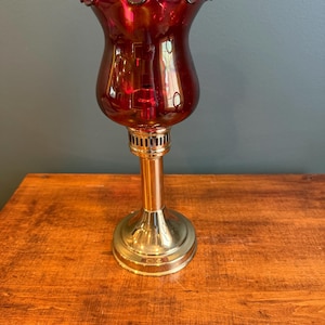 May include: A vintage ruby red glass lamp with a ruffled top. The lamp has a brass base and a copper-colored stem. The lamp is sitting on a wooden surface.