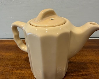 May include: Cream-colored ceramic teapot with a matching lid. The teapot has a faceted body, a curved handle, and a spout. The lid has a small handle and a hole. The teapot is sitting on a wooden surface.