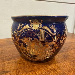 Könnte beinhalten: Ein runder, dunkelblauer Keramik-Blumentopf mit einem goldenen, verzierten Design. Das goldene Design zeigt Figuren, Bäume und architektonische Elemente. Der Rand des Topfes ist ebenfalls goldfarben.