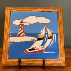 Puede incluir: Azulejo enmarcado con una escena náutica. La imagen muestra un velero con velas blancas y casco rojo, un faro con rayas rojas y blancas, y nubes blancas sobre un cielo y mar azules. El marco es de madera.