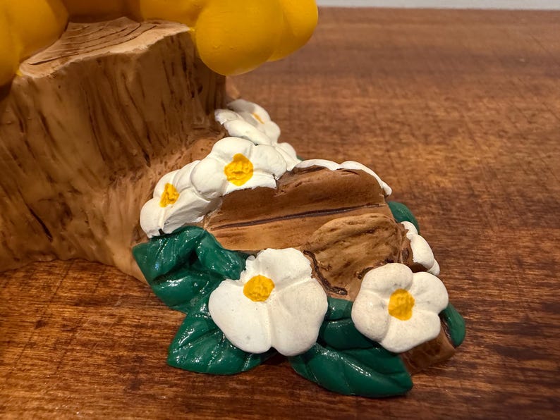 K&ouml;nnte beinhalten: Nahaufnahme eines dekorativen Figurenfu&szlig;es mit einer strukturierten, braunen, baumartigen Struktur, die mit wei&szlig;en Blumen mit gelben Zentren und gr&uuml;nen Bl&auml;ttern verziert ist. Der Sockel steht auf einer Holzoberfl&auml;che.