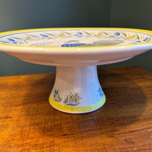 May include: A white ceramic cake stand with a yellow rim and a rooster design. The stand has a pedestal base and is decorated with blue and yellow floral accents. The cake stand is on a wooden surface.