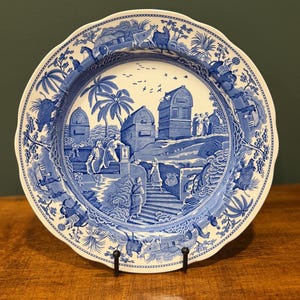 Peut inclure: Une assiette décorative bleue et blanche avec un bord festonné. L'assiette présente une scène de paysage détaillée avec des bâtiments, des personnages et des arbres. L'assiette est présentée sur un support en métal noir, mettant en valeur son design complexe et son esthétique vintage.