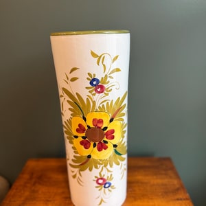 May include: A tall, cylindrical white ceramic vase featuring a hand-painted floral design. The pattern includes a large central yellow flower with red and brown accents, surrounded by green leaves, vines, and smaller blue and red floral motifs.