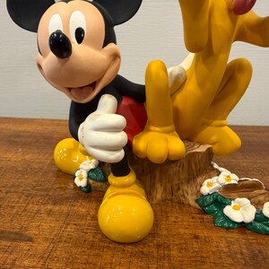 K&ouml;nnte beinhalten: Eine Figur mit Micky Maus und Pluto. Micky Maus kniet und hebt den Daumen. Pluto sitzt neben Micky. Die Figur steht auf einem Holzsockel mit wei&szlig;en Blumen.