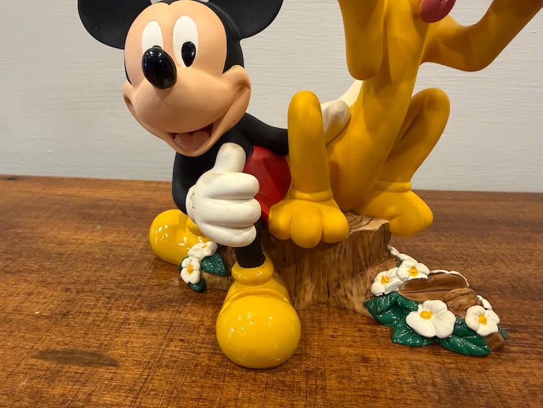 K&ouml;nnte beinhalten: Eine Figur mit Micky Maus und Pluto. Micky Maus kniet und hebt den Daumen. Pluto sitzt neben Micky. Die Figur steht auf einem Holzsockel mit wei&szlig;en Blumen.