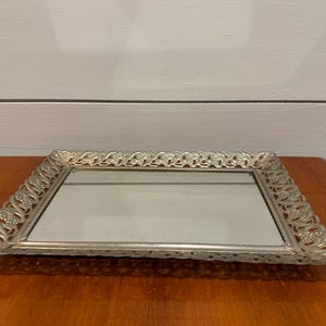 May include: A rectangular silver-toned vanity tray with a mirrored surface. The tray has a decorative, ornate border with floral and scrollwork details. The tray is displayed on a wooden surface.
