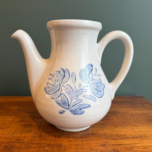 May include: Cream-colored ceramic pitcher with a spout and handle. The pitcher features blue floral designs on the front. The pitcher is sitting on a wooden surface.