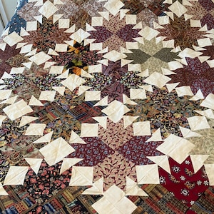 May include: A handmade quilt featuring a starburst pattern. The quilt showcases a variety of patterned fabrics in shades of burgundy, brown, blue, and green, with white star points. The quilt is a cozy bedding item.