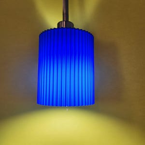 Puede incluir: Una lámpara colgante cilíndrica azul con una pantalla acanalada. La lámpara cuelga de una varilla plateada. La pantalla tiene un diseño plisado texturizado y emite un brillo azul brillante. El fondo es amarillo apagado.