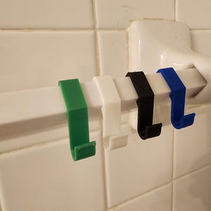 May include: Four 3D printed hooks in green, white, black, and blue colours are attached to a white metal bar. The hooks are designed to hang items on a shower rod or towel rack.