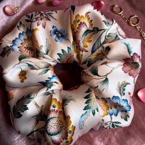 May include: A large, floral patterned scrunchie in cream, featuring a repeating design of blue, yellow, and pink flowers with green leaves. The fabric is gathered and knotted, creating a voluminous hair accessory. The scrunchie is a hair accessory.