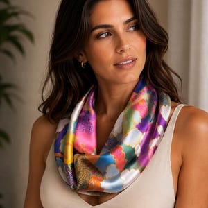 May include: A smooth, lustrous infinity scarf featuring a vibrant, multi-coloured abstract pattern with prominent shades of magenta, royal blue, bright yellow, and forest green, worn around the neck over a simple, solid-coloured beige tank top.