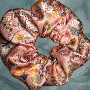 Op de afbeelding: Een roze satijnen scrunchie met een bloemenpatroon in tinten geel, groen, blauw en bruin.
