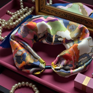 Può includere: Una fascia per capelli in seta colorata con un design paesaggistico nei toni del rosa, arancione, verde e viola. La fascia ha un nappa blu ed è esposta su una superficie rosa, con una collana di perle e un piccolo specchio sullo sfondo.