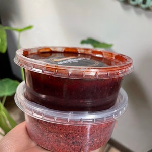 May include: Two clear plastic containers with lids, one stacked on top of the other. The top container is filled with a dark red sauce, while the bottom container is filled with a red powder.