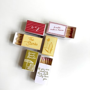 Peut inclure: Cinq boîtes d'allumettes rectangulaires avec divers motifs et textes. L'une présente des rayures rouges et 'C + J', d'autres portent les inscriptions 'A match made in Heaven', 'The Starks', 'Jensen Beach, Florida' avec une chaise de plage, et 'If you are reading this, Jack + Caty love you'.